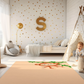 Kinderteppich "Abu Affe" von spielzwerg. Ambientebild von einem hellen Kinderzimmer mit weißem Holzboden und weißen Wänden, dekoriert mit goldenen Dreiecken und einem großen goldenen Buchstaben 'S'. Links steht ein gemütlicher Sessel mit Kissen und Decke, daneben eine goldene Stehlampe. In der Mitte liegt der Teppich. Rechts sitzt ein lachender Junge in einem weißen Spielzelt mit goldenen Punkten. Im Hintergrund ein schlichtes Bett mit weißer Decke und goldenen Akzenten. RUG-00128-XL, RUG-00128-L.