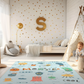 Kinderteppich "Monster" von spielzwerg. Ambientebild von einem hellen Kinderzimmer mit weißem Holzboden und weißen Wänden, dekoriert mit goldenen Dreiecken und einem großen goldenen Buchstaben 'S'. Links steht ein gemütlicher Sessel mit Kissen und Decke, daneben eine goldene Stehlampe. In der Mitte liegt der Teppich. Rechts sitzt ein lachender Junge in einem weißen Spielzelt mit goldenen Punkten. Im Hintergrund ein schlichtes Bett mit weißer Decke und goldenen Akzenten. RUG-00019-XL, RUG-00019-L.
