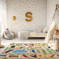 Spielteppich "Mosaik-Dorf" von spielzwerg. Ambientebild von einem hellen Kinderzimmer mit weißem Holzboden und weißen Wänden, dekoriert mit goldenen Dreiecken und einem großen goldenen Buchstaben 'S'. Links steht ein gemütlicher Sessel mit Kissen und Decke, daneben eine goldene Stehlampe. In der Mitte liegt der Teppich. Rechts sitzt ein lachender Junge in einem weißen Spielzelt mit goldenen Punkten. Im Hintergrund ein schlichtes Bett mit weißer Decke und goldenen Akzenten. RUG-00187-XL, RUG-00187-L.