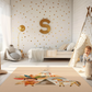 Kinderteppich "Indianer" von spielzwerg. Ambientebild von einem hellen Kinderzimmer mit weißem Holzboden und weißen Wänden, dekoriert mit goldenen Dreiecken und einem großen goldenen Buchstaben 'S'. Links steht ein gemütlicher Sessel mit Kissen und Decke, daneben eine goldene Stehlampe. In der Mitte liegt der Teppich. Rechts sitzt ein lachender Junge in einem weißen Spielzelt mit goldenen Punkten. Im Hintergrund ein schlichtes Bett mit weißer Decke und goldenen Akzenten. RUG-00186-XL, RUG-00186-L.