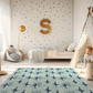 Kinderteppich "Orca Familie" von spielzwerg. Ambientebild von einem hellen Kinderzimmer mit weißem Holzboden und weißen Wänden, dekoriert mit goldenen Dreiecken und einem großen goldenen Buchstaben 'S'. Links steht ein gemütlicher Sessel mit Kissen und Decke, daneben eine goldene Stehlampe. In der Mitte liegt der Teppich. Rechts sitzt ein lachender Junge in einem weißen Spielzelt mit goldenen Punkten. Im Hintergrund ein schlichtes Bett mit weißer Decke und goldenen Akzenten. RUG-00102-XL, RUG-00102-L.