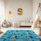 Kinderteppich "Hafenchaos" von spielzwerg. Ambientebild von einem hellen Kinderzimmer mit weißem Holzboden und weißen Wänden, dekoriert mit goldenen Dreiecken und einem großen goldenen Buchstaben 'S'. Links steht ein gemütlicher Sessel mit Kissen und Decke, daneben eine goldene Stehlampe. In der Mitte liegt der Teppich. Rechts sitzt ein lachender Junge in einem weißen Spielzelt mit goldenen Punkten. Im Hintergrund ein schlichtes Bett mit weißer Decke und goldenen Akzenten. RUG-00103-XL, RUG-00103-L.