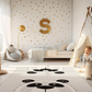 Kinderteppich "Boho Pandas" von spielzwerg. Ambientebild von einem hellen Kinderzimmer mit weißem Holzboden und weißen Wänden, dekoriert mit goldenen Dreiecken und einem großen goldenen Buchstaben 'S'. Links steht ein gemütlicher Sessel mit Kissen und Decke, daneben eine goldene Stehlampe. In der Mitte liegt der Teppich. Rechts sitzt ein lachender Junge in einem weißen Spielzelt mit goldenen Punkten. Im Hintergrund ein schlichtes Bett mit weißer Decke und goldenen Akzenten. RUG-00018-XL, RUG-00018-L.