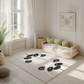 Kinderteppich "Boho Pandas" von spielzwerg. Ambientebild von einem hellen, freundlichen Kinderzimmer mit weißen Holzwänden und hellem Holzboden. In der Ecke steht ein niedriges Holzbett mit weißen Decken und hellgrünen Kissen. Davor liegt der Teppich. Links steht ein kleines Holzspielzeugrad am Fenster mit weißen Vorhängen. An der Wand hängt ein gerahmtes Bild mit einem dezenten, kreisförmigen Muster. RUG-00018-XL, RUG-00018-L.