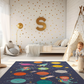 Kinderteppich "Sonnensystem mit Astronauten" von spielzwerg. Ambientebild von einem hellen Kinderzimmer mit weißem Holzboden und weißen Wänden, dekoriert mit goldenen Dreiecken und einem großen goldenen Buchstaben 'S'. Links steht ein gemütlicher Sessel mit Kissen und Decke, daneben eine goldene Stehlampe. In der Mitte liegt der Teppich. Rechts sitzt ein lachender Junge in einem weißen Spielzelt mit goldenen Punkten. Im Hintergrund ein schlichtes Bett mit weißer Decke und goldenen Akzenten. RUG-00101-XL, RU