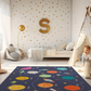 Kinderteppich "Sonnensystem" von spielzwerg. Ambientebild von einem hellen Kinderzimmer mit weißem Holzboden und weißen Wänden, dekoriert mit goldenen Dreiecken und einem großen goldenen Buchstaben 'S'. Links steht ein gemütlicher Sessel mit Kissen und Decke, daneben eine goldene Stehlampe. In der Mitte liegt der Teppich. Rechts sitzt ein lachender Junge in einem weißen Spielzelt mit goldenen Punkten. Im Hintergrund ein schlichtes Bett mit weißer Decke und goldenen Akzenten. RUG-00143-XL, RUG-00143-L.