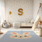 Kinderteppich "Hasi" von spielzwerg. Ambientebild von einem hellen Kinderzimmer mit weißem Holzboden und weißen Wänden, dekoriert mit goldenen Dreiecken und einem großen goldenen Buchstaben 'S'. Links steht ein gemütlicher Sessel mit Kissen und Decke, daneben eine goldene Stehlampe. In der Mitte liegt der Teppich. Rechts sitzt ein lachender Junge in einem weißen Spielzelt mit goldenen Punkten. Im Hintergrund ein schlichtes Bett mit weißer Decke und goldenen Akzenten. RUG-00129-XL, RUG-00129-L.