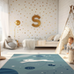 Kinderteppich "Hase im All" von spielzwerg. Ambientebild von einem hellen Kinderzimmer mit weißem Holzboden und weißen Wänden, dekoriert mit goldenen Dreiecken und einem großen goldenen Buchstaben 'S'. Links steht ein gemütlicher Sessel mit Kissen und Decke, daneben eine goldene Stehlampe. In der Mitte liegt der Teppich. Rechts sitzt ein lachender Junge in einem weißen Spielzelt mit goldenen Punkten. Im Hintergrund ein schlichtes Bett mit weißer Decke und goldenen Akzenten. RUG-00001-XL, RUG-00001-L.