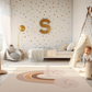 Kinderteppich "Regenbogen Twins" von spielzwerg. Ambientebild von einem hellen Kinderzimmer mit weißem Holzboden und weißen Wänden, dekoriert mit goldenen Dreiecken und einem großen goldenen Buchstaben 'S'. Links steht ein gemütlicher Sessel mit Kissen und Decke, daneben eine goldene Stehlampe. In der Mitte liegt der Teppich. Rechts sitzt ein lachender Junge in einem weißen Spielzelt mit goldenen Punkten. Im Hintergrund ein schlichtes Bett mit weißer Decke und goldenen Akzenten. RUG-00184-XL, RUG-00184-L.