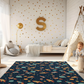 Kinderteppich „Weltraumchaos“ von spielzwerg. Ambientebild von einem hellen Kinderzimmer mit weißem Holzboden und weißen Wänden, dekoriert mit goldenen Dreiecken und einem großen goldenen Buchstaben 'S'. Links steht ein gemütlicher Sessel mit Kissen und Decke, daneben eine goldene Stehlampe. In der Mitte liegt der Teppich. Rechts sitzt ein lachender Junge in einem weißen Spielzelt mit goldenen Punkten. Im Hintergrund ein schlichtes Bett mit weißer Decke und goldenen Akzenten. RUG-00155-XL, RUG-00155-L.