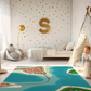 Spielteppich "Stadt am Meer" von spielzwerg. Ambientebild von einem hellen Kinderzimmer mit weißem Holzboden und weißen Wänden, dekoriert mit goldenen Dreiecken und einem großen goldenen Buchstaben 'S'. Links steht ein gemütlicher Sessel mit Kissen und Decke, daneben eine goldene Stehlampe. In der Mitte liegt der Teppich. Rechts sitzt ein lachender Junge in einem weißen Spielzelt mit goldenen Punkten. Im Hintergrund ein schlichtes Bett mit weißer Decke und goldenen Akzenten. RUG-00040-XL, RUG-00040-L.
