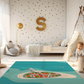 Spielteppich "Inseldorf" von spielzwerg. Ambientebild von einem hellen Kinderzimmer mit weißem Holzboden und weißen Wänden, dekoriert mit goldenen Dreiecken und einem großen goldenen Buchstaben 'S'. Links steht ein gemütlicher Sessel mit Kissen und Decke, daneben eine goldene Stehlampe. In der Mitte liegt der Teppich. Rechts sitzt ein lachender Junge in einem weißen Spielzelt mit goldenen Punkten. Im Hintergrund ein schlichtes Bett mit weißer Decke und goldenen Akzenten. RUG-00039-XL, RUG-00039-L.