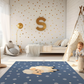 Kinderteppich "Samu Schäfchen" von spielzwerg. Ambientebild von einem hellen Kinderzimmer mit weißem Holzboden und weißen Wänden, dekoriert mit goldenen Dreiecken und einem großen goldenen Buchstaben 'S'. Links steht ein gemütlicher Sessel mit Kissen und Decke, daneben eine goldene Stehlampe. In der Mitte liegt der Teppich. Rechts sitzt ein lachender Junge in einem weißen Spielzelt mit goldenen Punkten. Im Hintergrund ein schlichtes Bett mit weißer Decke und goldenen Akzenten. RUG-00148-XL, RUG-00148-L.