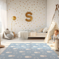 Kinderteppich "Flauschige Wolken" von spielzwerg. Ambientebild von einem hellen Kinderzimmer mit weißem Holzboden und weißen Wänden, dekoriert mit goldenen Dreiecken und einem großen goldenen Buchstaben 'S'. Links steht ein gemütlicher Sessel mit Kissen und Decke, daneben eine goldene Stehlampe. In der Mitte liegt der Teppich. Rechts sitzt ein lachender Junge in einem weißen Spielzelt mit goldenen Punkten. Im Hintergrund ein schlichtes Bett mit weißer Decke und goldenen Akzenten. RUG-00016-XL, RUG-00016-L.
