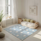 Kinderteppich "Flauschige Wolken" von spielzwerg. Ambientebild von einem hellen, freundlichen Kinderzimmer mit weißen Holzwänden und hellem Holzboden. In der Ecke steht ein niedriges Holzbett mit weißen Decken und hellgrünen Kissen. Davor liegt der Teppich. Links steht ein kleines Holzspielzeugrad am Fenster mit weißen Vorhängen. An der Wand hängt ein gerahmtes Bild mit einem dezenten, kreisförmigen Muster. RUG-00016-XL, RUG-00016-L.