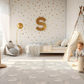 Kinderteppich "Weiße Schäfchen" von spielzwerg. Ambientebild von einem hellen Kinderzimmer mit weißem Holzboden und weißen Wänden, dekoriert mit goldenen Dreiecken und einem großen goldenen Buchstaben 'S'. Links steht ein gemütlicher Sessel mit Kissen und Decke, daneben eine goldene Stehlampe. In der Mitte liegt der Teppich. Rechts sitzt ein lachender Junge in einem weißen Spielzelt mit goldenen Punkten. Im Hintergrund ein schlichtes Bett mit weißer Decke und goldenen Akzenten. RUG-00015-XL, RUG-00015-L.