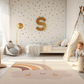 Kinderteppich "Regenbogen Twin" von spielzwerg. Ambientebild von einem hellen Kinderzimmer mit weißem Holzboden und weißen Wänden, dekoriert mit goldenen Dreiecken und einem großen goldenen Buchstaben 'S'. Links steht ein gemütlicher Sessel mit Kissen und Decke, daneben eine goldene Stehlampe. In der Mitte liegt der Teppich. Rechts sitzt ein lachender Junge in einem weißen Spielzelt mit goldenen Punkten. Im Hintergrund ein schlichtes Bett mit weißer Decke und goldenen Akzenten. RUG-00182-XL, RUG-00182-L.