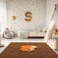 Kinderteppich "Finn Fuchs" von spielzwerg. Ambientebild von einem hellen Kinderzimmer mit weißem Holzboden und weißen Wänden, dekoriert mit goldenen Dreiecken und einem großen goldenen Buchstaben 'S'. Links steht ein gemütlicher Sessel mit Kissen und Decke, daneben eine goldene Stehlampe. In der Mitte liegt der Teppich. Rechts sitzt ein lachender Junge in einem weißen Spielzelt mit goldenen Punkten. Im Hintergrund ein schlichtes Bett mit weißer Decke und goldenen Akzenten. RUG-00149-XL, RUG-00149-L.