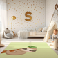 Kinderteppich "Flori Faultier" von spielzwerg. Ambientebild von einem hellen Kinderzimmer mit weißem Holzboden und weißen Wänden, dekoriert mit goldenen Dreiecken und einem großen goldenen Buchstaben 'S'. Links steht ein gemütlicher Sessel mit Kissen und Decke, daneben eine goldene Stehlampe. In der Mitte liegt der Teppich. Rechts sitzt ein lachender Junge in einem weißen Spielzelt mit goldenen Punkten. Im Hintergrund ein schlichtes Bett mit weißer Decke und goldenen Akzenten. RUG-00135-XL, RUG-00135-L.