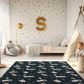 Kinderteppich "Weltraumbären" von spielzwerg. Ambientebild von einem hellen Kinderzimmer mit weißem Holzboden und weißen Wänden, dekoriert mit goldenen Dreiecken und einem großen goldenen Buchstaben 'S'. Links steht ein gemütlicher Sessel mit Kissen und Decke, daneben eine goldene Stehlampe. In der Mitte liegt der Teppich. Rechts sitzt ein lachender Junge in einem weißen Spielzelt mit goldenen Punkten. Im Hintergrund ein schlichtes Bett mit weißer Decke und goldenen Akzenten. RUG-00136-XL, RUG-00136-L.