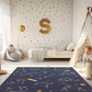 Kinderteppich "Sternzeichen" von spielzwerg. Ambientebild von einem hellen Kinderzimmer mit weißem Holzboden und weißen Wänden, dekoriert mit goldenen Dreiecken und einem großen goldenen Buchstaben 'S'. Links steht ein gemütlicher Sessel mit Kissen und Decke, daneben eine goldene Stehlampe. In der Mitte liegt der Teppich. Rechts sitzt ein lachender Junge in einem weißen Spielzelt mit goldenen Punkten. Im Hintergrund ein schlichtes Bett mit weißer Decke und goldenen Akzenten. RUG-00099-XL, RUG-00099-L.