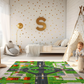 Spielteppich "Große Kreuzung" von spielzwerg. Ambientebild von einem hellen Kinderzimmer mit weißem Holzboden und weißen Wänden, dekoriert mit goldenen Dreiecken und einem großen goldenen Buchstaben 'S'. Links steht ein gemütlicher Sessel mit Kissen und Decke, daneben eine goldene Stehlampe. In der Mitte liegt der Teppich. Rechts sitzt ein lachender Junge in einem weißen Spielzelt mit goldenen Punkten. Im Hintergrund ein schlichtes Bett mit weißer Decke und goldenen Akzenten. RUG-00035-XL, RUG-00035-L.