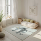 Kinderteppich "Boho Katze" von spielzwerg. Ambientebild von einem hellen, freundlichen Kinderzimmer mit weißen Holzwänden und hellem Holzboden. In der Ecke steht ein niedriges Holzbett mit weißen Decken und hellgrünen Kissen. Davor liegt der Teppich. Links steht ein kleines Holzspielzeugrad am Fenster mit weißen Vorhängen. An der Wand hängt ein gerahmtes Bild mit einem dezenten, kreisförmigen Muster. RUG-00038-XL, RUG-00038-L.