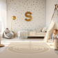 Spielteppich "Zielscheibe" von spielzwerg. Ambientebild von einem hellen Kinderzimmer mit weißem Holzboden und weißen Wänden, dekoriert mit goldenen Dreiecken und einem großen goldenen Buchstaben 'S'. Links steht ein gemütlicher Sessel mit Kissen und Decke, daneben eine goldene Stehlampe. In der Mitte liegt der Teppich. Rechts sitzt ein lachender Junge in einem weißen Spielzelt mit goldenen Punkten. Im Hintergrund ein schlichtes Bett mit weißer Decke und goldenen Akzenten. RUG-00178-XL, RUG-00178-L.