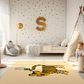 Kinderteppich "Tiger mit Blumen" von spielzwerg. Ambientebild von einem hellen Kinderzimmer mit weißem Holzboden und weißen Wänden, dekoriert mit goldenen Dreiecken und einem großen goldenen Buchstaben 'S'. Links steht ein gemütlicher Sessel mit Kissen und Decke, daneben eine goldene Stehlampe. In der Mitte liegt der Teppich. Rechts sitzt ein lachender Junge in einem weißen Spielzelt mit goldenen Punkten. Im Hintergrund ein schlichtes Bett mit weißer Decke und goldenen Akzenten. RUG-00122-XL, RUG-00122-L.