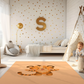Kinderteppich "Tigerstark" von spielzwerg. Ambientebild von einem hellen Kinderzimmer mit weißem Holzboden und weißen Wänden, dekoriert mit goldenen Dreiecken und einem großen goldenen Buchstaben 'S'. Links steht ein gemütlicher Sessel mit Kissen und Decke, daneben eine goldene Stehlampe. In der Mitte liegt der Teppich. Rechts sitzt ein lachender Junge in einem weißen Spielzelt mit goldenen Punkten. Im Hintergrund ein schlichtes Bett mit weißer Decke und goldenen Akzenten. RUG-00013-XL, RUG-00013-L.