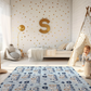 Kinderteppich „Tier-Lokomotive“ von spielzwerg. Ambientebild von einem hellen Kinderzimmer mit weißem Holzboden und weißen Wänden, dekoriert mit goldenen Dreiecken und einem großen goldenen Buchstaben 'S'. Links steht ein gemütlicher Sessel mit Kissen und Decke, daneben eine goldene Stehlampe. In der Mitte liegt der Teppich. Rechts sitzt ein lachender Junge in einem weißen Spielzelt mit goldenen Punkten. Im Hintergrund ein schlichtes Bett mit weißer Decke und goldenen Akzenten. RUG-00074-XL, RUG-00074-L.
