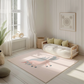 Kinderteppich "Emmy Einhorn" von spielzwerg. Ambientebild von einem hellen, freundlichen Kinderzimmer mit weißen Holzwänden und hellem Holzboden. In der Ecke steht ein niedriges Holzbett mit weißen Decken und hellgrünen Kissen. Davor liegt der Teppich. Links steht ein kleines Holzspielzeugrad am Fenster mit weißen Vorhängen. An der Wand hängt ein gerahmtes Bild mit einem dezenten, kreisförmigen Muster. RUG-00117-XL, RUG-00117-L.