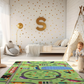 Spielteppich "Dorf am Waldrand" von spielzwerg. Ambientebild von einem hellen Kinderzimmer mit weißem Holzboden und weißen Wänden, dekoriert mit goldenen Dreiecken und einem großen goldenen Buchstaben 'S'. Links steht ein gemütlicher Sessel mit Kissen und Decke, daneben eine goldene Stehlampe. In der Mitte liegt der Teppich. Rechts sitzt ein lachender Junge in einem weißen Spielzelt mit goldenen Punkten. Im Hintergrund ein schlichtes Bett mit weißer Decke und goldenen Akzenten. RUG-00012-XL, RUG-00012-L.