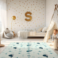 Kinderteppich "Karo Wale" von spielzwerg. Ambientebild von einem hellen Kinderzimmer mit weißem Holzboden und weißen Wänden, dekoriert mit goldenen Dreiecken und einem großen goldenen Buchstaben 'S'. Links steht ein gemütlicher Sessel mit Kissen und Decke, daneben eine goldene Stehlampe. In der Mitte liegt der Teppich. Rechts sitzt ein lachender Junge in einem weißen Spielzelt mit goldenen Punkten. Im Hintergrund ein schlichtes Bett mit weißer Decke und goldenen Akzenten. RUG-00139-XL, RUG-00139-L.