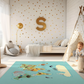 Spielteppich "Tiere der Welt" von spielzwerg. Ambientebild von einem hellen Kinderzimmer mit weißem Holzboden und weißen Wänden, dekoriert mit goldenen Dreiecken und einem großen goldenen Buchstaben 'S'. Links steht ein gemütlicher Sessel mit Kissen und Decke, daneben eine goldene Stehlampe. In der Mitte liegt der Teppich. Rechts sitzt ein lachender Junge in einem weißen Spielzelt mit goldenen Punkten. Im Hintergrund ein schlichtes Bett mit weißer Decke und goldenen Akzenten. RUG-00177-XL, RUG-00177-L.
