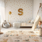 Spielteppich "Niedlicher Zoo" von spielzwerg. Ambientebild von einem hellen Kinderzimmer mit weißem Holzboden und weißen Wänden, dekoriert mit goldenen Dreiecken und einem großen goldenen Buchstaben 'S'. Links steht ein gemütlicher Sessel mit Kissen und Decke, daneben eine goldene Stehlampe. In der Mitte liegt der Teppich. Rechts sitzt ein lachender Junge in einem weißen Spielzelt mit goldenen Punkten. Im Hintergrund ein schlichtes Bett mit weißer Decke und goldenen Akzenten. RUG-00011-XL, RUG-00011-L.